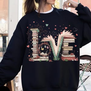 May include: Black sweatshirt with the word "LOVE" spelled out with stacks of books. The "L" and "E" are made of books, and the "O" is a wreath with books and a quill. Pink and green hearts are scattered around the design.