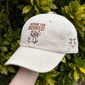May include: A beige baseball cap with the text "PREPARE FOR BEEYATLE!" and an embroidered cartoon goat holding a potion. The cap also features a goat head emblem on the side. The hat is made of a soft, durable fabric.