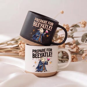 May include: Two ceramic mugs, one black and one white, stacked. Each mug features the phrase "PREPARE FOR BEEYATLE!" and an illustration of a goat in a wizard robe holding a potion. The mugs have a tan base and a curved handle.