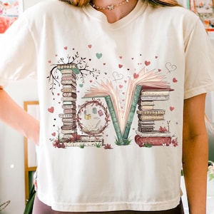 May include: Cream-colored t-shirt featuring the word "LOVE" formed by stacks of books and an open book. The "L" and "E" are made of stacked books, while the "O" is a wreath of books. Hearts and floral accents complete the design.