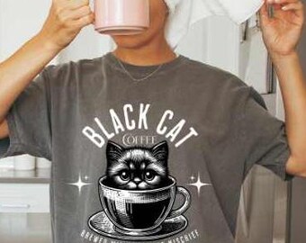 May include: A gray t-shirt with a graphic of a black cat in a coffee cup, the text reads "Black Cat Coffee" and "Brewed with a side of mischief". The person is holding a pink mug. Pink flowers are in a vase.