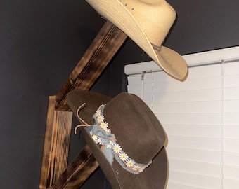 May include: A wooden hat rack with two cowboy hats. The top hat is light brown and the bottom hat is dark brown with a white daisy trim.
