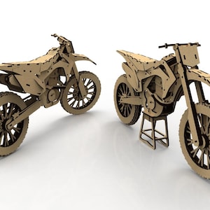 May include: Two cardboard model dirt bikes. The bikes are made of light brown cardboard with dark brown accents. One bike is on a stand. The tires have a detailed tread pattern. The bikes are on a white surface.