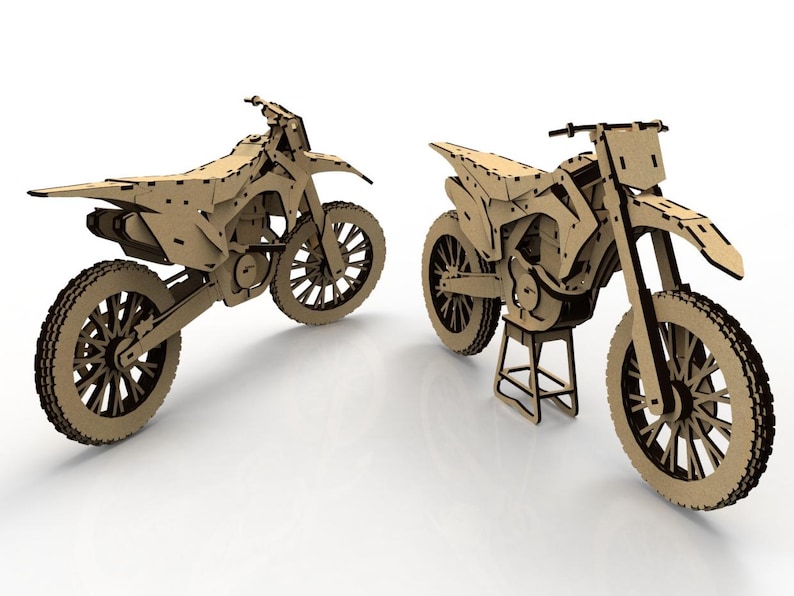 May include: Two cardboard model dirt bikes. The bikes are made of light brown cardboard with dark brown accents. One bike is on a stand. The tires have a detailed tread pattern. The bikes are on a white surface.