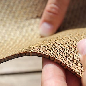 May include: Close-up of a flexible, woven material with a repeating geometric pattern. The material is a light brown color, and the pattern is made up of small, square-shaped designs. The material is being held by a person's fingers.