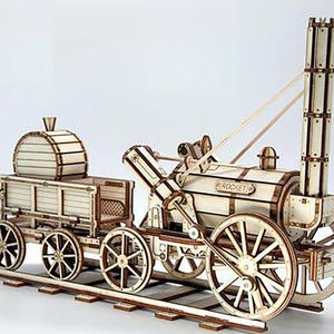 May include: A wooden model of a vintage steam train, featuring a locomotive and a separate car. The model is constructed from light-colored wood with visible details like wheels, a smokestack, and the word "ROCKET" on the side.