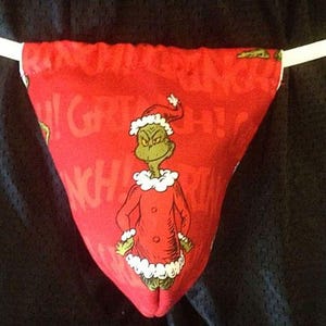 May include: Red thong underwear featuring a cartoon Grinch character in a Santa hat and coat. The Grinch is green with a white collar and cuffs. The word "Grinch" is printed on the red fabric.