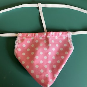 May include: A pink fabric pouch with white polka dots and white lace trim. The pouch is attached to a white elastic band.