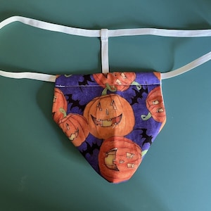 May include: Purple fabric with a white elastic band. The fabric has a pattern of orange jack-o'-lantern faces on a purple background.