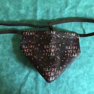 May include: Black bandana with "HAPPY NEW YEAR" text and colorful confetti print. The bandana has a black elastic band attached for wearing. The bandana is made of fabric.