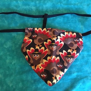 May include: A black fabric triangle with a black elastic band. The fabric has a pattern of brown turkeys with red, yellow, and white feathers.