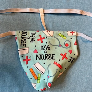 Op de afbeelding: Een lichtblauwe stoffen tas met een witte elastische band. De tas heeft een herhalend patroon van rode kruisen, harten, stethoscopen en de tekst "Love a Nurse".