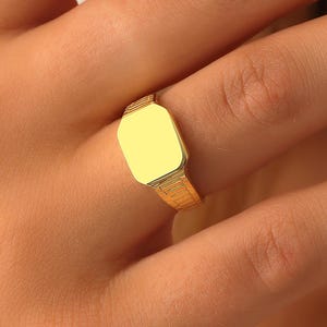 K&ouml;nnte beinhalten: Ein goldener Siegelring mit einer glatten, flachen, rechteckigen Oberfl&auml;che und abgeschr&auml;gten Kanten. Die Ringschiene weist ein texturiertes, geripptes Muster auf, das sich von der Mitte aus &uuml;ber die Seiten erstreckt.