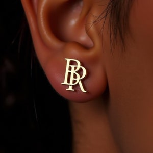 May include: A pair of gold initial earrings with the letters "BR" intertwined. The earrings are shown in a close-up view on a person's ear.