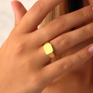 K&ouml;nnte beinhalten: Ein goldener Siegelring mit einer glatten, flachen, rechteckigen Oberfl&auml;che und abgeschr&auml;gten Kanten. Die Ringschiene weist ein texturiertes, geripptes Muster auf, das sich von der Mitte aus &uuml;ber die Seiten erstreckt.