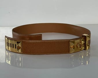 Pode incluir: Um cinto de couro castanho claro com ferragens metálicas douradas. O cinto apresenta uma fivela retangular com ranhuras verticais e um fecho decorativo com um anel e tachas piramidais. O cinto é exibido numa superfície refletora.