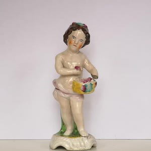 May include: A porcelain figurine of a young person with brown hair and a pink dress holding a basket of fruit. The figurine is painted with pink, green, and yellow colors.
