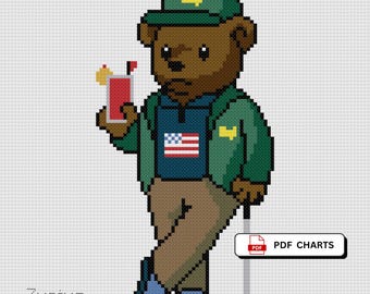 May include: Cross-stitch design of a brown bear wearing a green cap and jacket, holding a red drink with a garnish. The bear is dressed in golf attire, with a golf club and the American flag on its shirt. The text "PDF CHARTS" is visible.