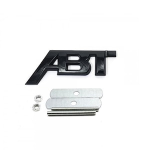 Op de afbeelding: Zwart plastic "ABT" embleem met twee zilveren montageplaten en schroeven.