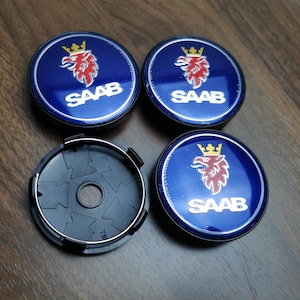 Könnte beinhalten: Vier blaue und silberne Saab-Radnabenabdeckungen. Jede Kappe zeigt das Saab-Logo: einen roten Greif mit goldener Krone und den Schriftzug "SAAB" in Weiß. Eine schwarze Kunststoffkappe ist ebenfalls sichtbar.