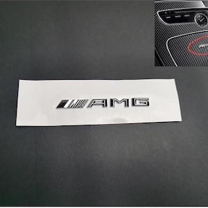 Puede incluir: Un emblema AMG plateado y negro sobre un respaldo blanco, con un primer plano del interior de un coche que muestra el mismo emblema. El emblema es un accesorio para el coche.