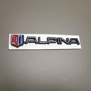 Può includere: Emblema nero "ALPINA" con un logo a scudo con dettagli rossi, bianchi e blu. L'emblema è montato su una base rettangolare bianca, su sfondo grigio.