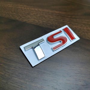 Peut inclure: Un emblème TSI chromé et rouge sur un support blanc. Les lettres T et I sont chromées, tandis que le S est rouge avec un contour chromé. L'emblème est posé sur une surface à grain de bois foncé.