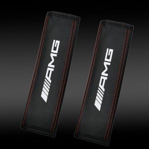 Puede incluir: Dos fundas negras para cinturones de seguridad con las letras blancas "AMG" y costuras rojas. Las fundas son rectangulares y parecen estar hechas de cuero o un material similar. El fondo es un degradado de negro y gris.