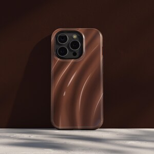 Può includere: Una custodia per telefono marrone con un motivo a spirale a forma di onda. La custodia ha una finitura lucida e un ritaglio per la fotocamera con tre obiettivi. Lo sfondo è marrone pieno e la custodia poggia su una superficie bianca.