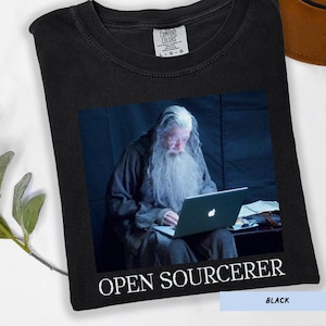 May include: Black t-shirt featuring a graphic of a wizard using a laptop, with the text "OPEN SOURCERER" below the image. The shirt is labeled "BLACK" and has a Comfort Colors tag.