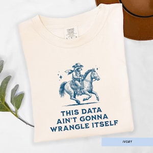 May include: Ivory-colored t-shirt with a blue illustration of a raccoon riding a horse, wearing a cowboy hat. The text "THIS DATA AIN'T GONNA WRANGLE ITSELF" is printed below the graphic. The shirt is folded.