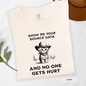 May include: Ivory-colored t-shirt with the text "SHOW ME YOUR SOURCE DATA AND NO ONE GETS HURT." The shirt features a graphic of a kitten wearing a cowboy hat and bandana, with cacti in the background.