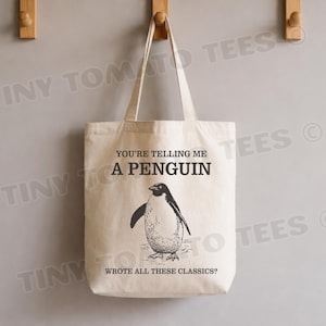 May include: Cream-colored tote bag with a penguin illustration and the text "YOU'RE TELLING ME A PENGUIN WROTE ALL THESE CLASSICS?". The penguin is black and white. The bag has two handles and is hanging on a wall.