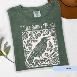 May include: Moss green t-shirt with the text "I Like Shiny Things" above a black raven illustration. The raven is surrounded by a floral design. The shirt is a Comfort Colors brand.