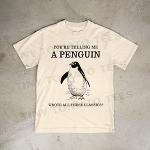 May include: Cream-colored t-shirt featuring a black and white illustration of a penguin. The text on the shirt reads, "YOU'RE TELLING ME A PENGUIN WROTE ALL THESE CLASSICS?" A humorous graphic tee.