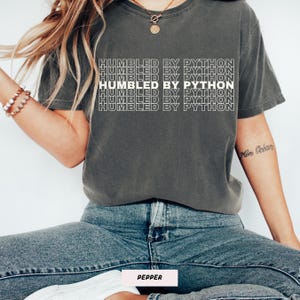 May include: A dark gray t-shirt with the text "HUMBLED BY PYTHON" in a stacked white font. The shirt is worn with blue jeans. The word "PEPPER" is at the bottom of the image.