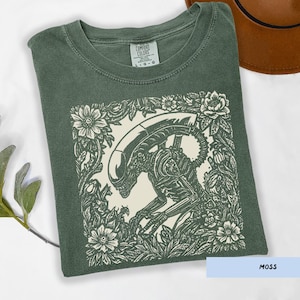 May include: Moss green t-shirt with a cream-colored graphic of an alien surrounded by floral elements. The shirt has a crew neck and short sleeves. The text "COMFORT COLORS" is visible on the tag.