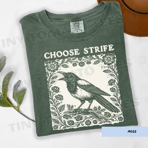 May include: Moss green t-shirt with the words "CHOOSE STRIFE" above a black and white illustration of a crow within a floral frame. The shirt is a Comfort Colors brand, size L-G-G.