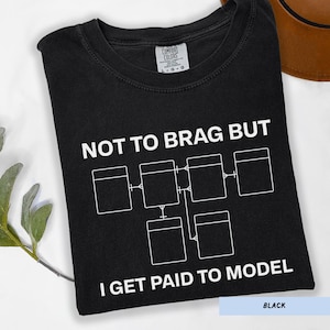 May include: Black t-shirt with white text and a diagram. The text reads "NOT TO BRAG BUT I GET PAID TO MODEL". The shirt has a crew neck and short sleeves. The shirt is folded and has a tag that says "COMFORT COLORS".