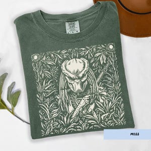 May include: A moss green t-shirt featuring a white graphic of a Predator character surrounded by a floral design. The shirt has a crew neck and short sleeves. The text "COMFORT COLORS" is visible on the upper left side of the shirt.