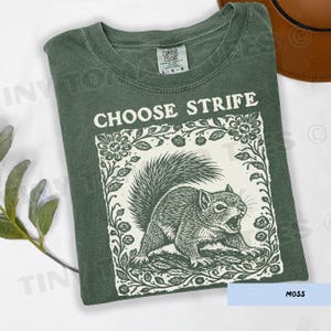 May include: Moss green t-shirt with the text "CHOOSE STRIFE" above a detailed illustration of a squirrel within a floral frame. The shirt is a Comfort Colors brand, size L-G.