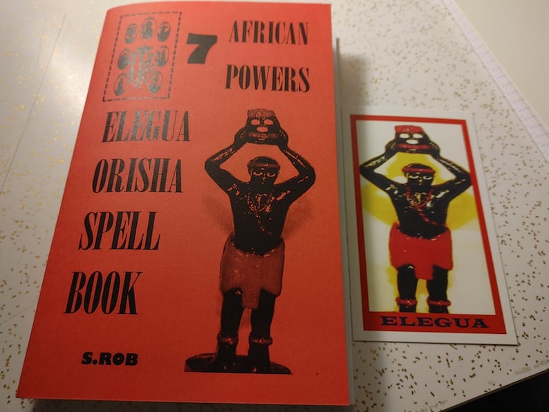 May include: A red book titled "Elegua Orisha Spell Book" by S. Rob. The book cover features a black and red illustration of a figure holding a pot. The book is next to a card with a similar illustration and the text "Elegua" on it.