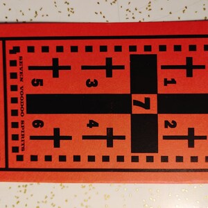 Op de afbeelding: Een rode kaart met een zwart raster en kruisen. De tekst "SEVEN VOODOO SPIRITS" en "RETURN LOVER OR SPOUSE" staat op de kaart gedrukt.