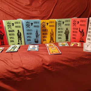Pode incluir: Sete livros coloridos intitulados "African Powers" com os nomes de diferentes divindades Orisha e as palavras "Orisha Spell Book" em cada capa. Os livros est&atilde;o dispostos em uma fila sobre uma superf&iacute;cie vermelha.