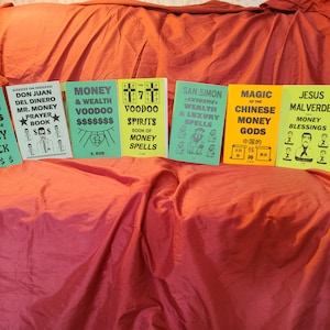 May include: A collection of nine small, green, yellow, and orange paperback books with black text. The books are titled "7 African Powers", "Don Juan Del Dinero", "Money & Wealth Voodoo", "Voodoo Spirits", "San Simon", "Magic of the Chinese Money Gods", "Jesus Malverde", and "Billionaire Voodoo Rituals".