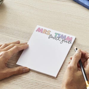 May include: A rectangular white notepad featuring the colourful text MRS TIMM in a playful font, with the words fourth grade written in black cursive script underneath. The notepad is positioned on a light wood grain surface.