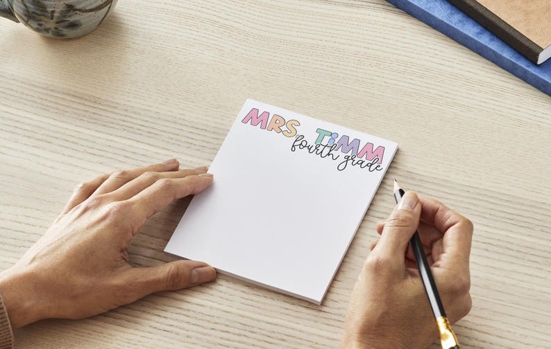 May include: A rectangular white notepad featuring the colourful text MRS TIMM in a playful font, with the words fourth grade written in black cursive script underneath. The notepad is positioned on a light wood grain surface.