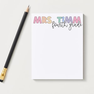 May include: A rectangular notepad with white paper featuring the text MRS. TIMM in colorful pastel block letters and the words fourth grade in black cursive script below it, accompanied by a black wooden pencil with a gold ferrule and eraser.