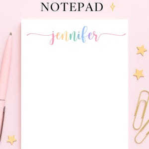 May include: A white personalized notepad with the name "Jennifer" in a colorful script font. The notepad is surrounded by a pink background, a pink pen, gold paper clips, and star-shaped decorations. The text "PERSONALIZED NOTEPAD" is at the top.