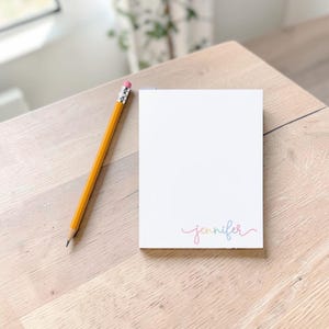 Pode incluir: Um bloco de notas retangular branco com o nome Jennifer escrito em letra cursiva colorida no canto inferior direito, acompanhado por um lápis de madeira amarelo com uma virola prateada e uma borracha rosa posicionado à esquerda.